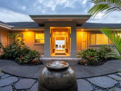 Nighttime view of the front entry courtyard featuring a central water vessel fountain and illuminated walkway leading to the entry doors. Landscape lighting highlights plants and architectural surfaces.