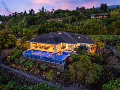 Elevated aerial perspective of a single-story residential property positioned on a landscaped lot with mature tropical vegetation. The home features a dark roofline, expansive covered lanai, and an ocean-facing infinity-edge swimming pool and spa. Lava rock garden walls and layered plantings surround the structure, with clear sightlines toward the Pacific Ocean and Kona coastline.